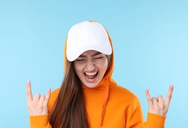 Photo of Happy woman in stylish baseball hat showing rock gesture on light blue background