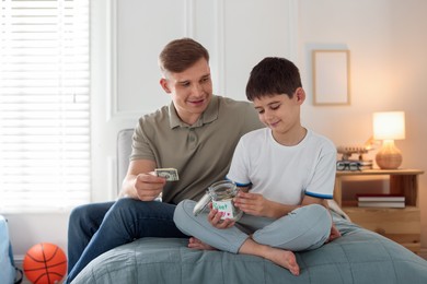 Photo of Father and son putting pocket money into jar on bed at home