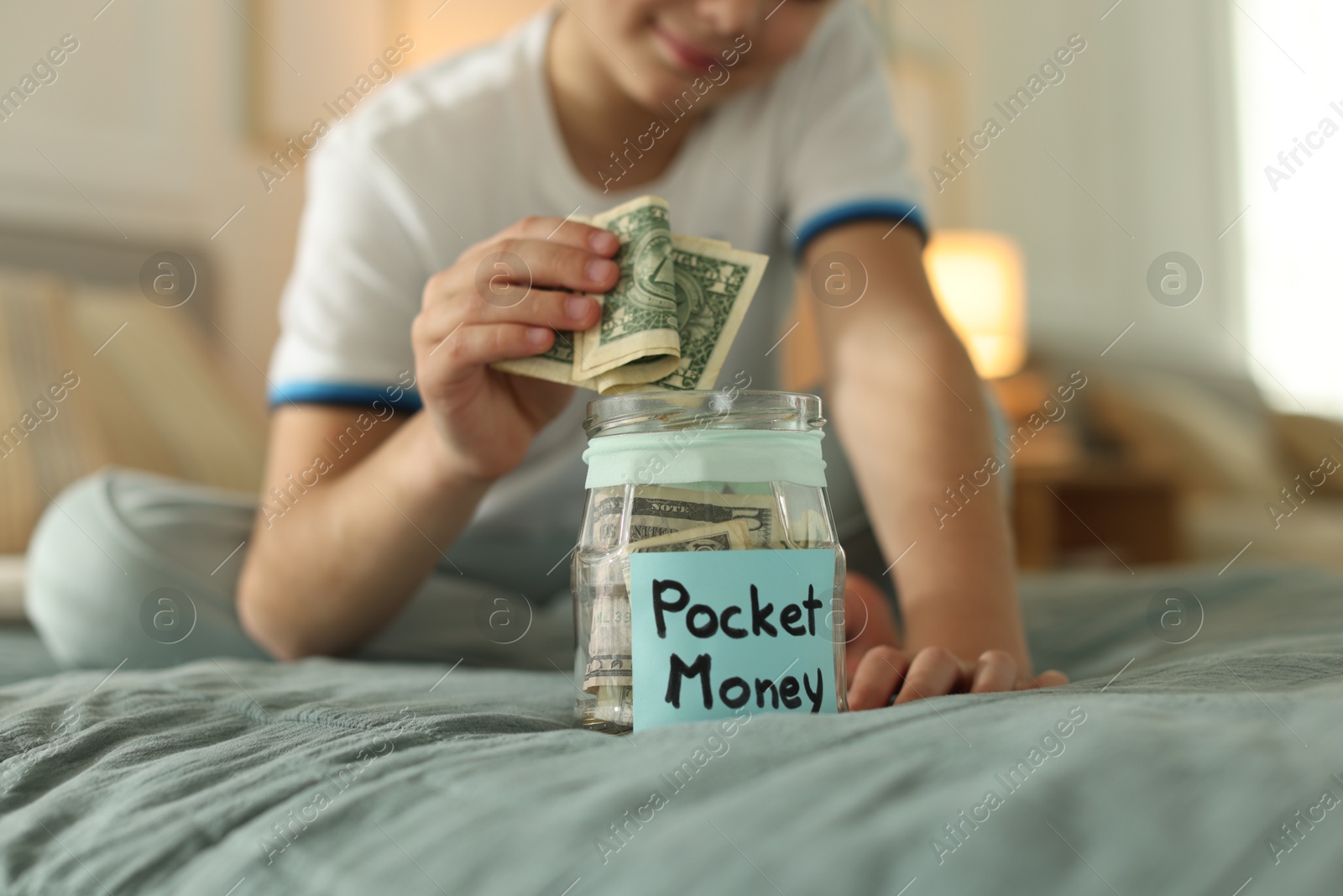 Boy putting pocket money into jar on bed at home, closeup Photo of Boy putting pocket money into jar on bed at home, closeup