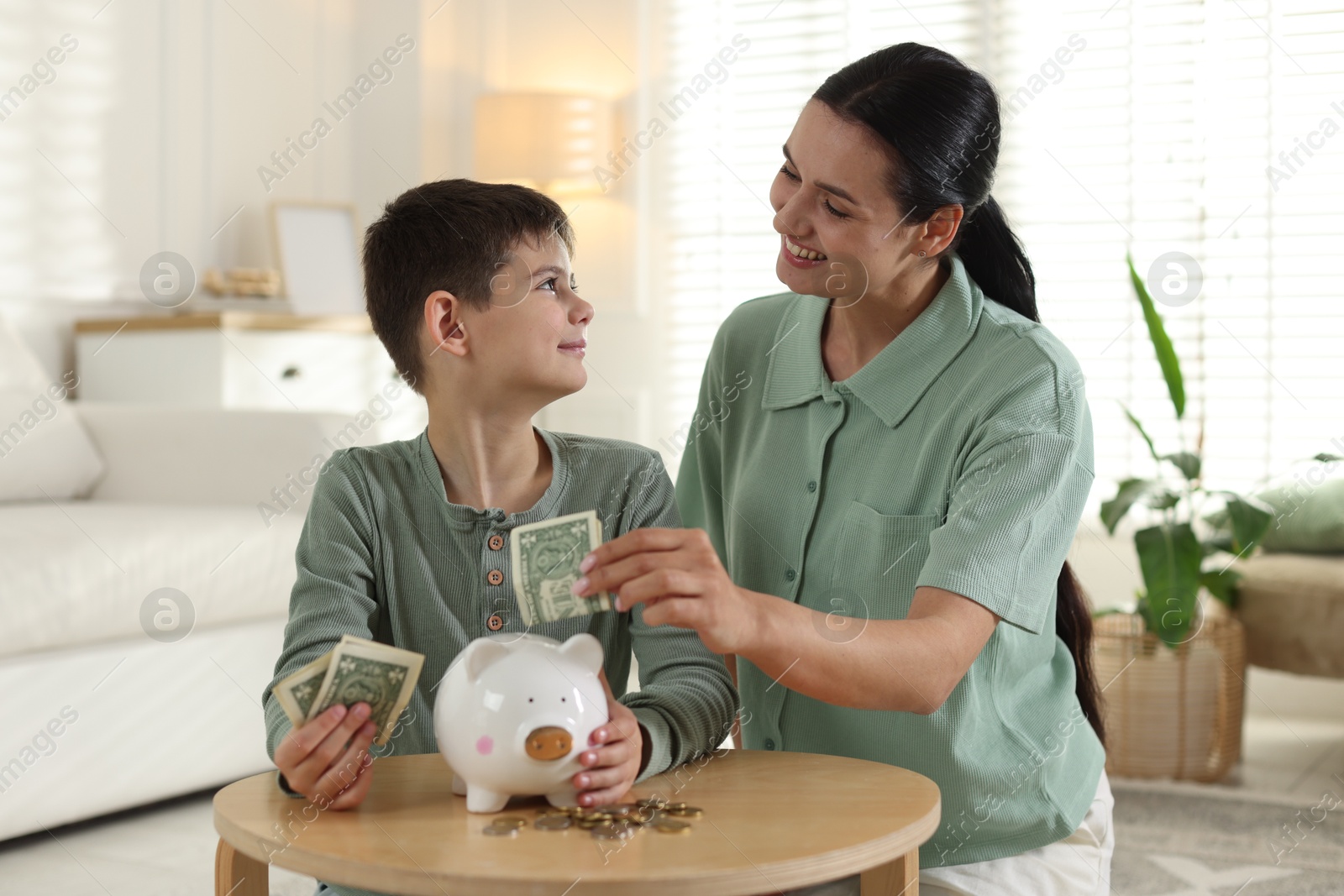 Mother and son putting pocket money into piggybank at table indoors Photo of Mother and son putting pocket money into piggybank at table indoors