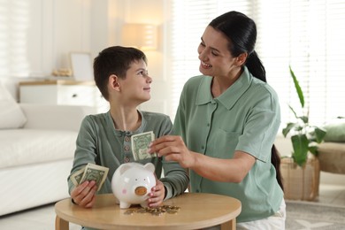 Photo of Mother and son putting pocket money into piggybank at table indoors