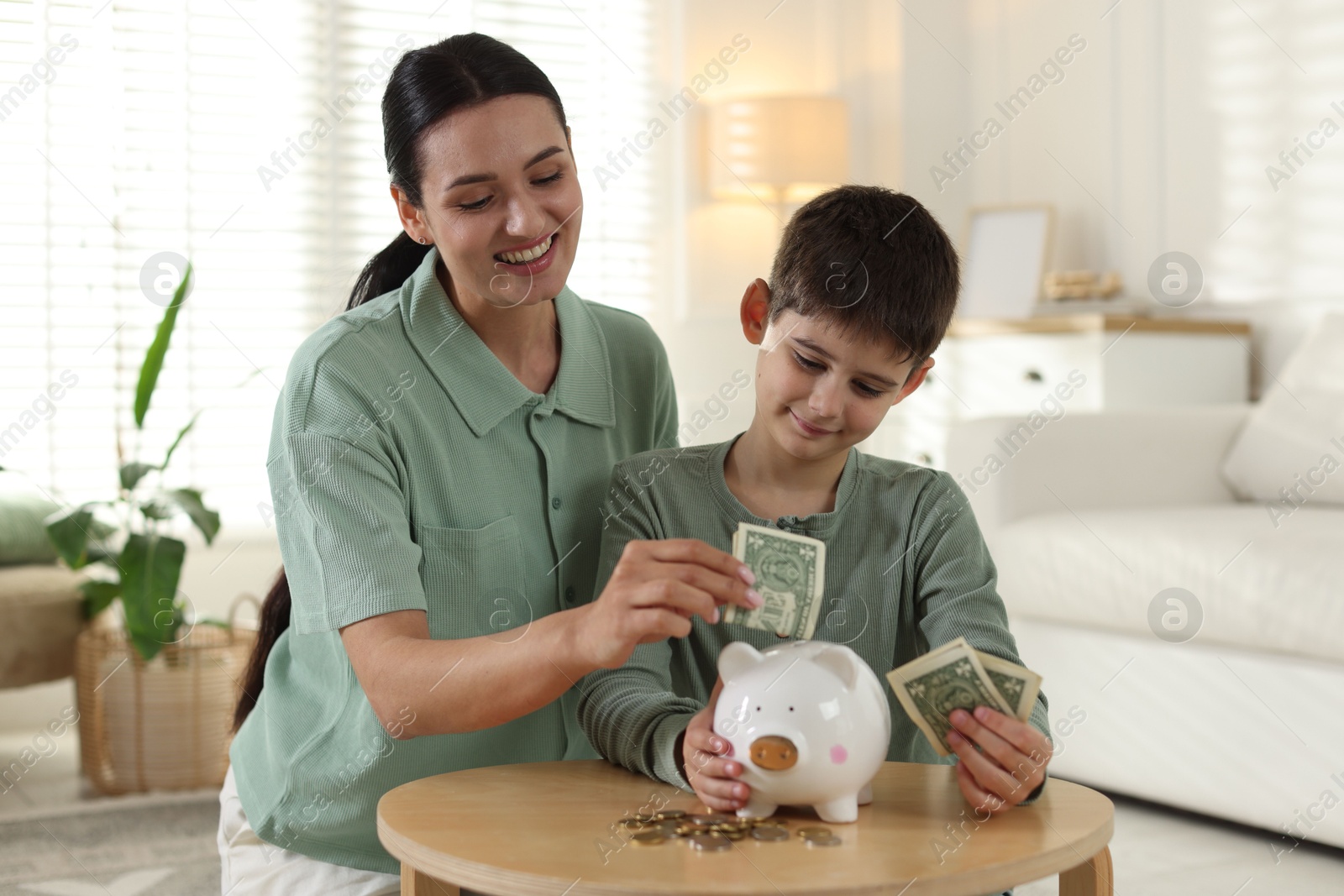 Mother and son putting pocket money into piggybank at table indoors Photo of Mother and son putting pocket money into piggybank at table indoors
