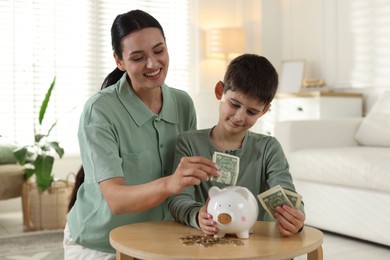 Photo of Mother and son putting pocket money into piggybank at table indoors