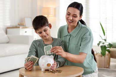 Photo of Mother and son putting pocket money into piggybank at table indoors