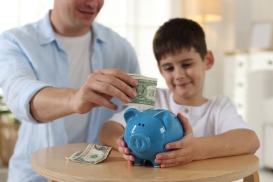 Photo of Father and son putting pocket money into piggybank at table indoors, selective focus