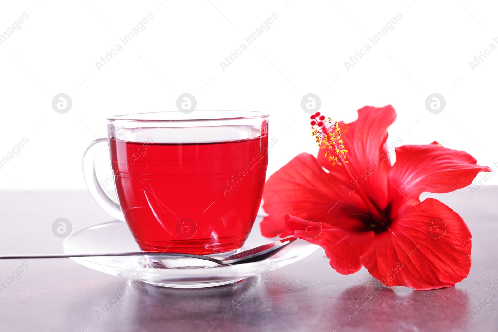 Tasty hibiscus tea in cup, spoon and beautiful flower on grey textured table against white background Photo of Tasty hibiscus tea in cup, spoon and beautiful flower on grey textured table against white background