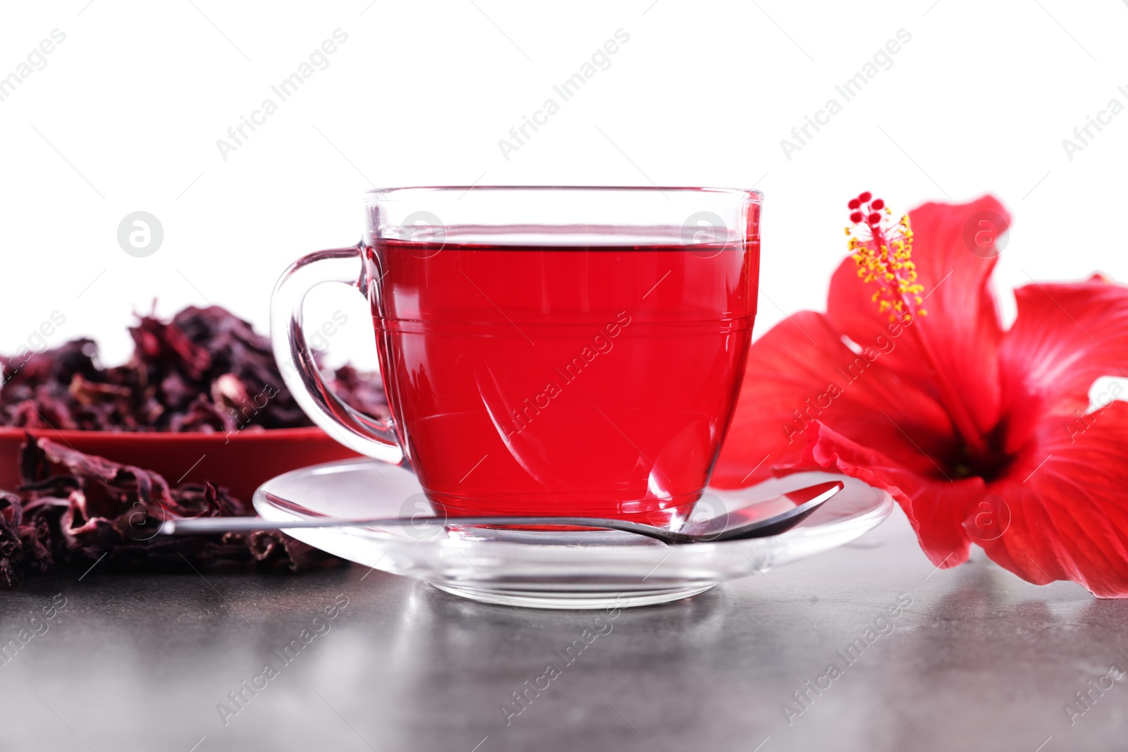Tasty hibiscus tea in cup, dry roselle sepals and beautiful flower on grey textured table against white background Photo of Tasty hibiscus tea in cup, dry roselle sepals and beautiful flower on grey textured table against white background