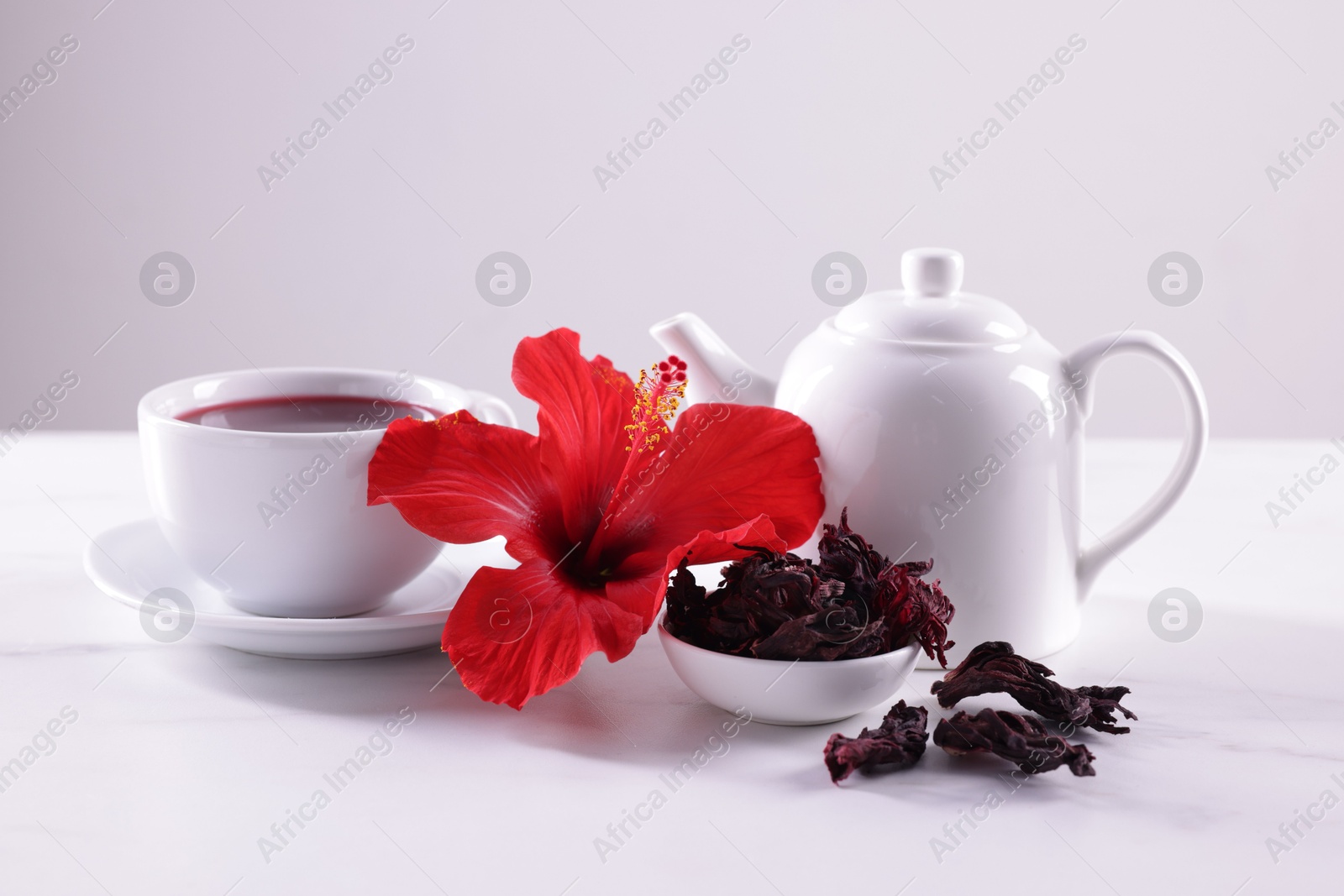 Tasty hibiscus tea, dry roselle sepals and beautiful flower on white table against grey background Photo of Tasty hibiscus tea, dry roselle sepals and beautiful flower on white table against grey background