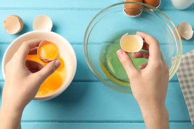 Photo of Woman separating egg yolk from white over glass bowl at light blue wooden table, top view