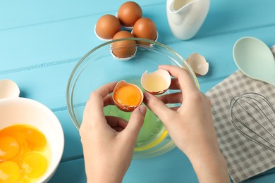 Photo of Woman separating egg yolk from white over glass bowl at light blue wooden table, above view