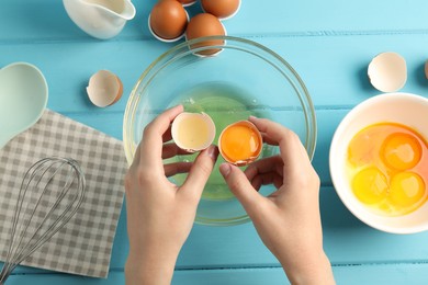 Photo of Woman separating egg yolk from white over glass bowl at light blue wooden table, top view