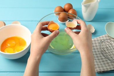 Photo of Woman separating egg yolk from white over glass bowl at light blue wooden table, closeup