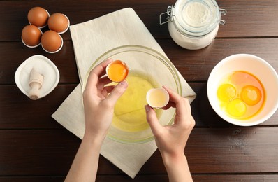 Photo of Woman separating egg yolk from white over glass bowl at wooden table, top view
