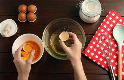 Photo of Woman separating egg yolk from white over glass bowl at wooden table, top view