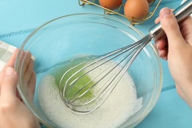 Photo of Woman whisking egg whites in bowl at light blue wooden table, closeup