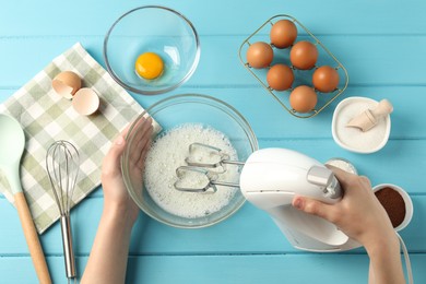 Photo of Woman whisking egg whites with hand mixer at light blue wooden table, top view