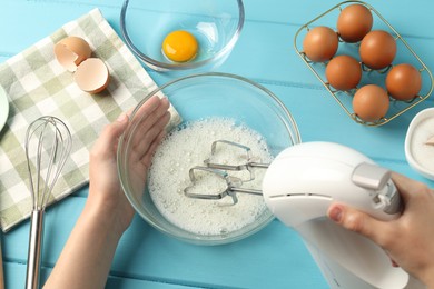 Photo of Woman whisking egg whites with hand mixer at light blue wooden table, above view