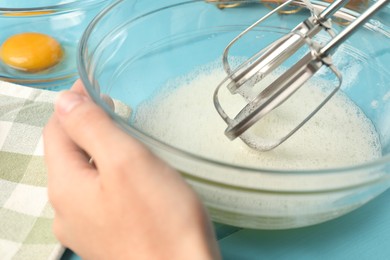 Photo of Woman whisking egg whites with hand mixer at light blue wooden table, closeup