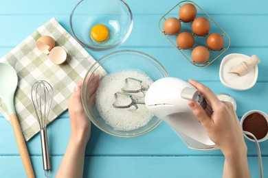 Photo of Woman whisking egg whites with hand mixer at light blue wooden table, top view