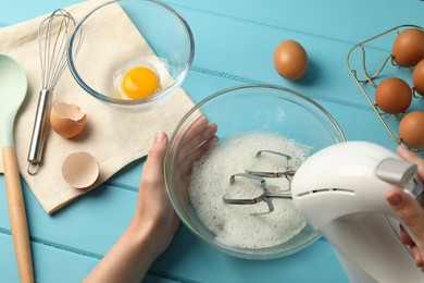 Photo of Woman whisking egg whites with hand mixer at light blue wooden table, above view