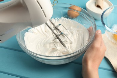 Photo of Woman whisking cream with hand mixer at light blue wooden table, closeup