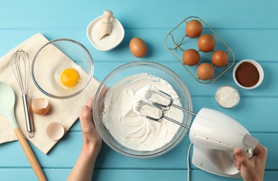 Photo of Woman whisking cream with hand mixer at light blue wooden table, top view