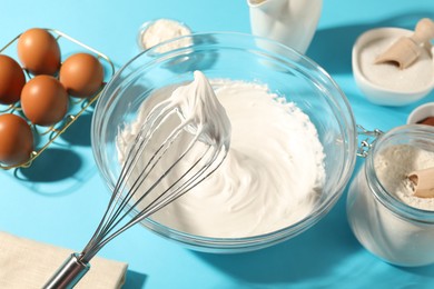 Photo of Whisk with whipped cream, eggs and other products on light blue table, closeup