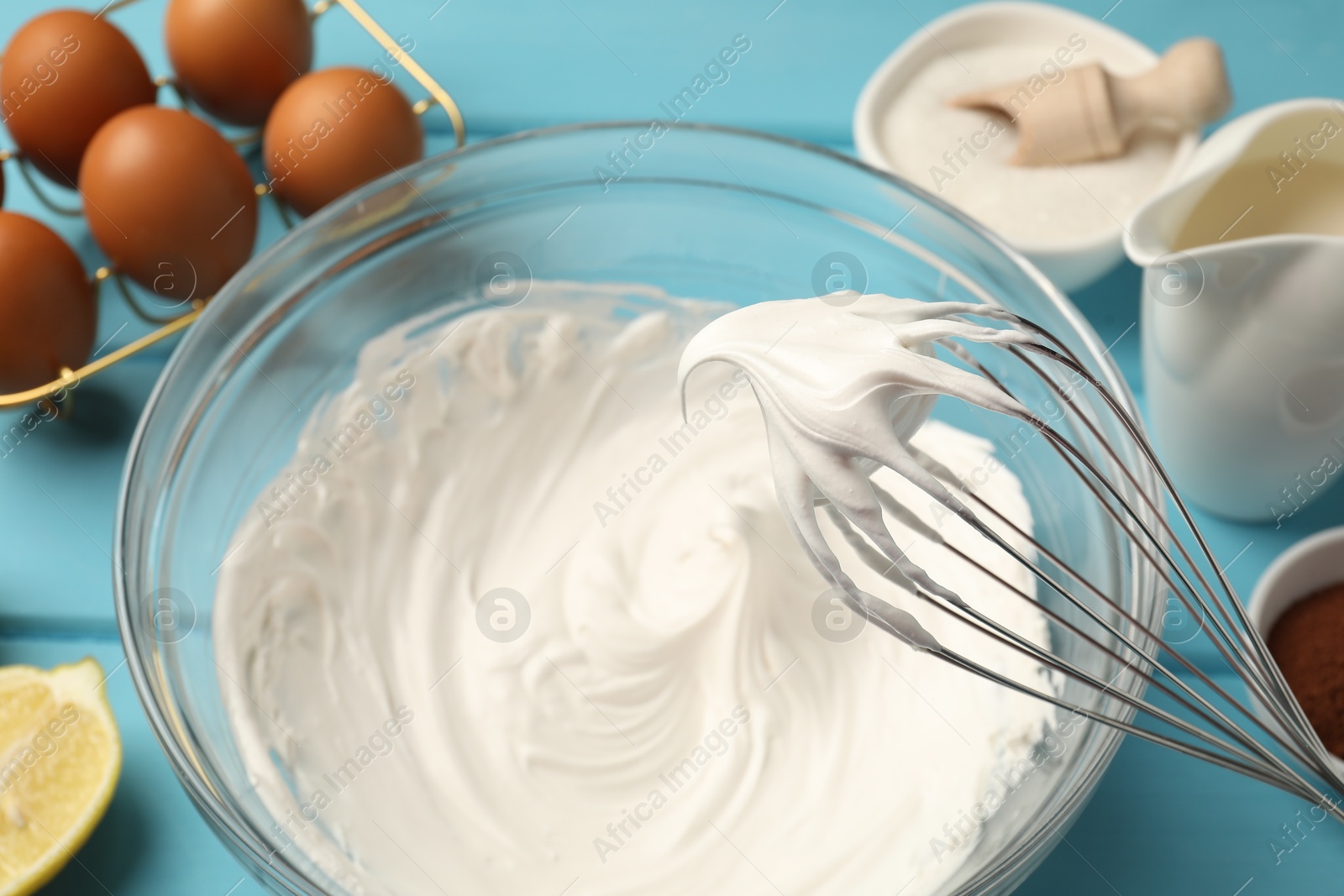 Whisk with whipped cream above bowl, eggs and other products on light blue table, closeup Photo of Whisk with whipped cream above bowl, eggs and other products on light blue table, closeup