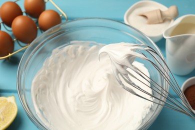 Photo of Whisk with whipped cream above bowl, eggs and other products on light blue table, closeup