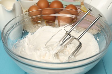 Photo of Hand mixer with whisks, whipped cream in bowl, eggs and milk on light blue table, closeup