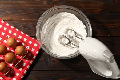 Photo of Hand mixer, whipped cream in bowl and eggs on wooden table, flat lay