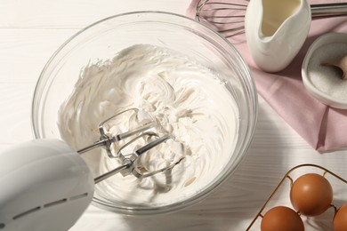 Photo of Hand mixer with whipped cream, eggs and other products on white wooden table, above view