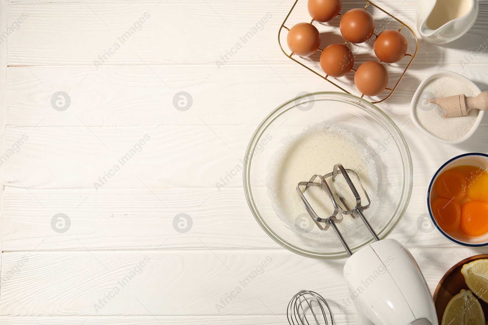 Hand mixer, egg whites in bowl, eggs and other products on white wooden table, flat lay. Space for text Photo of Hand mixer, egg whites in bowl, eggs and other products on white wooden table, flat lay. Space for text