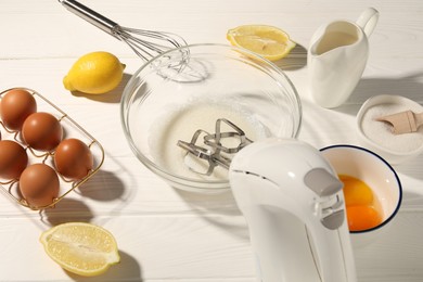 Photo of Hand mixer, egg whites in bowl, eggs and other products on white wooden table