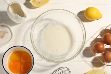 Photo of Whisk, egg whites in bowl, eggs and other products on white wooden table, flat lay