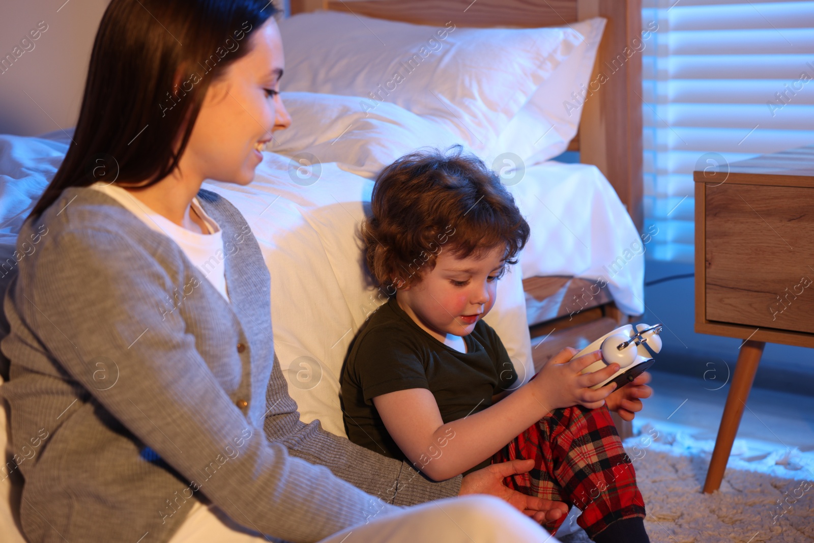 Happy mother and her son with alarm clock before bedtime in bedroom Photo of Happy mother and her son with alarm clock before bedtime in bedroom