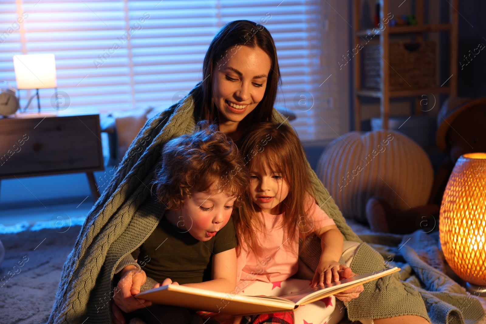 Smiling mother wrapped in blanket reading bedtime story to her children on floor at home Photo of Smiling mother wrapped in blanket reading bedtime story to her children on floor at home