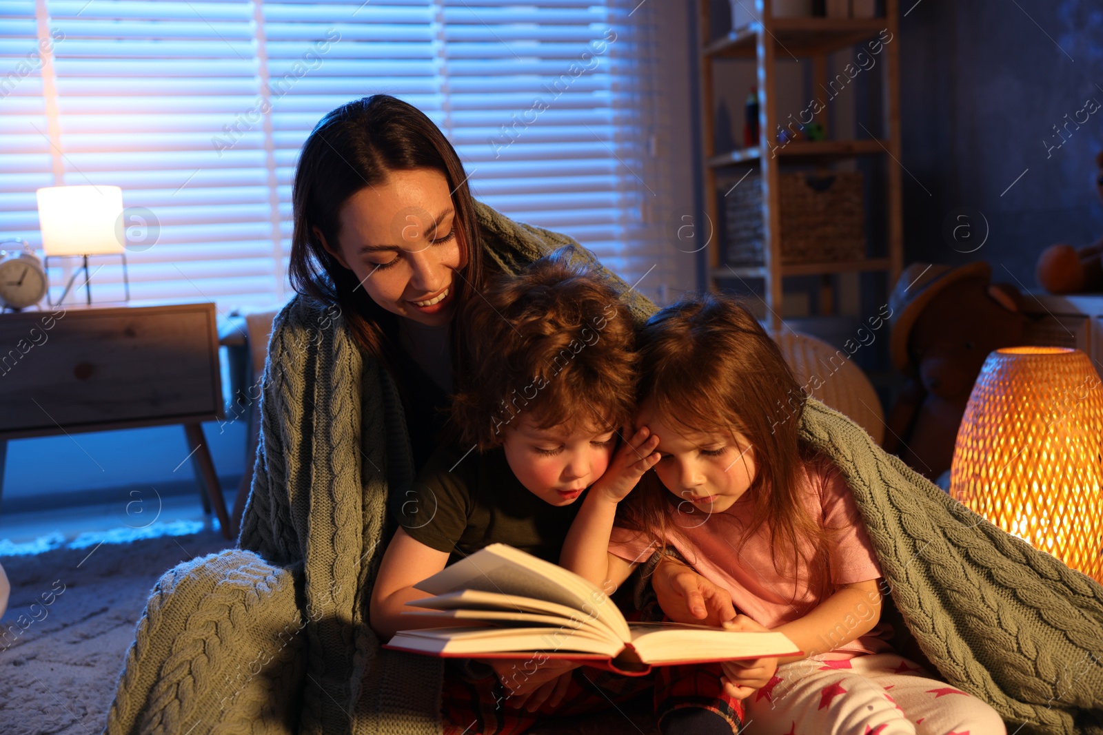 Smiling mother wrapped in blanket reading bedtime story to her children on floor at home Photo of Smiling mother wrapped in blanket reading bedtime story to her children on floor at home