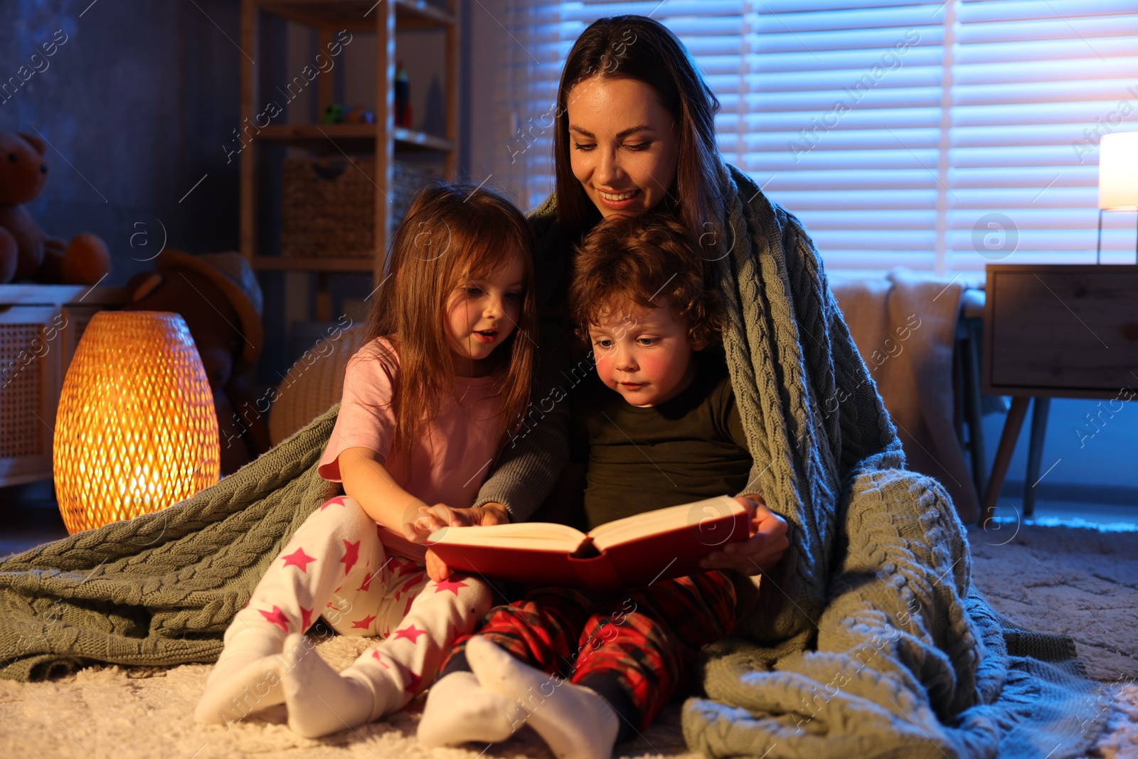 Smiling mother wrapped in blanket reading bedtime story to her children on floor at home Photo of Smiling mother wrapped in blanket reading bedtime story to her children on floor at home
