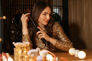 Photo of Woman curling her beautiful hair near mirror indoors