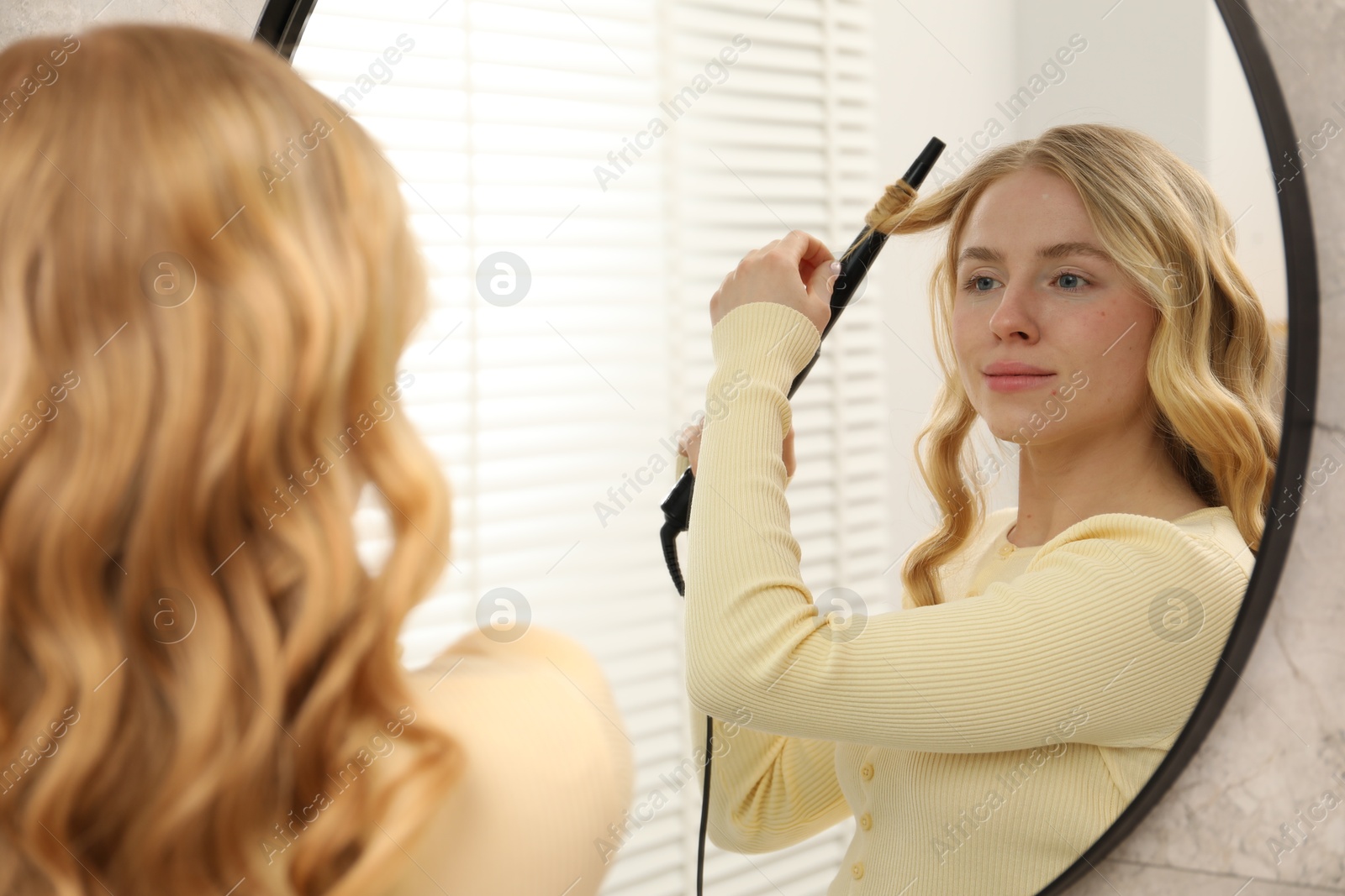 Woman curling her hair near mirror indoors Photo of Woman curling her hair near mirror indoors