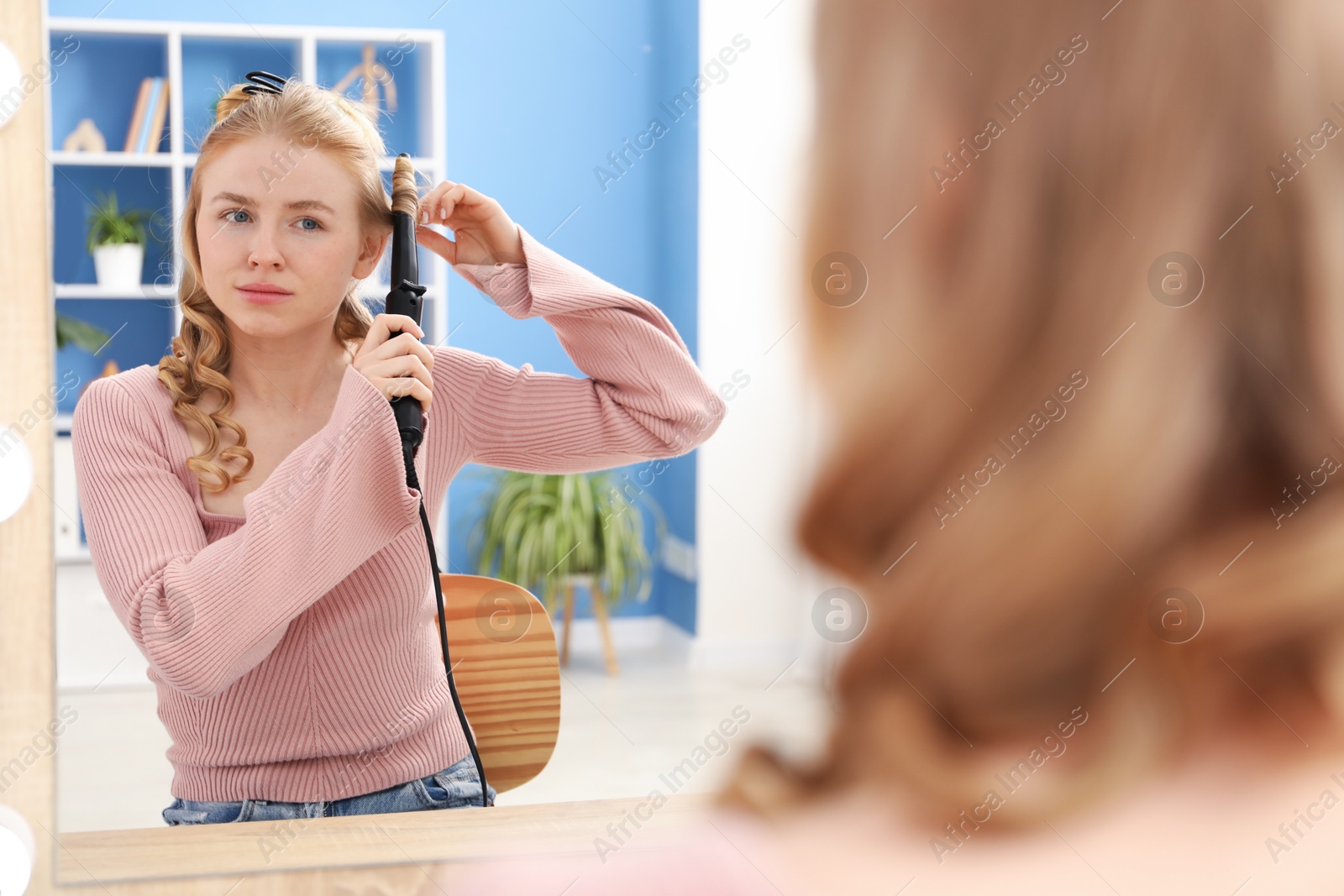 Woman curling her hair near mirror indoors Photo of Woman curling her hair near mirror indoors