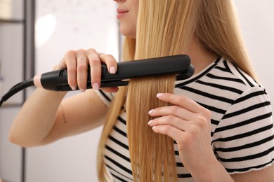 Photo of Woman straightening her beautiful long hair indoors, closeup