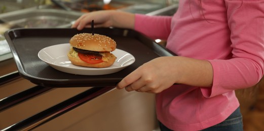 Photo of Little girl with plastic tray and burger near serving line in canteen, closeup. School food
