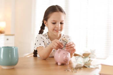 Photo of Girl putting pocket money into piggybank at table indoors