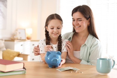 Photo of Mother and daughter putting pocket money into piggybank at table indoors