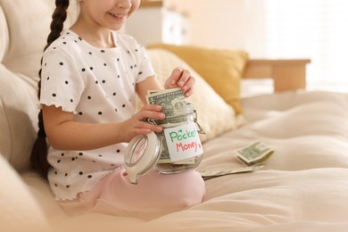 Photo of Girl putting her pocket money into jar on sofa at home, closeup