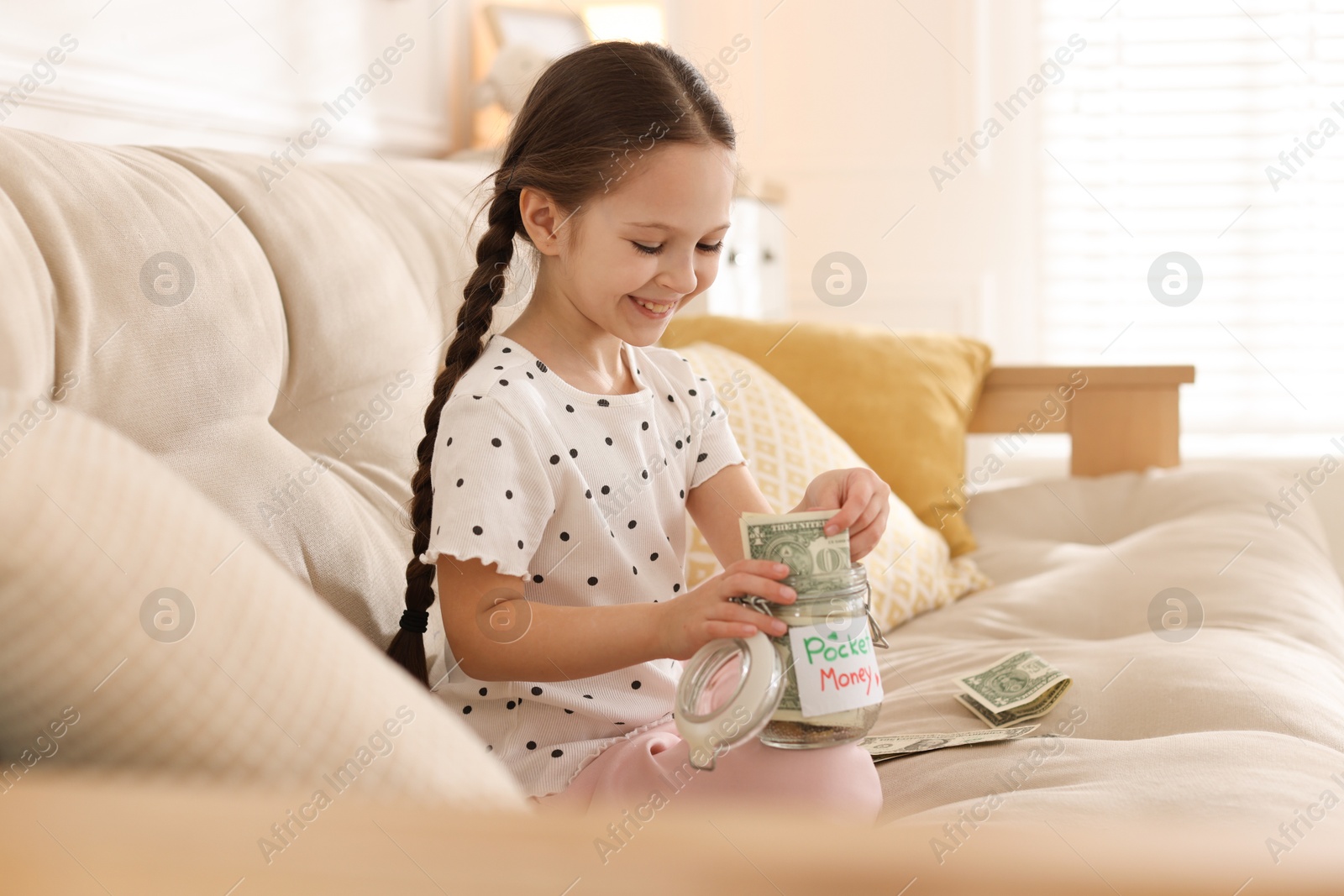 Girl putting her pocket money into jar on sofa at home Photo of Girl putting her pocket money into jar on sofa at home