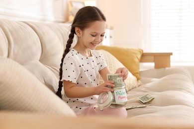 Photo of Girl putting her pocket money into jar on sofa at home