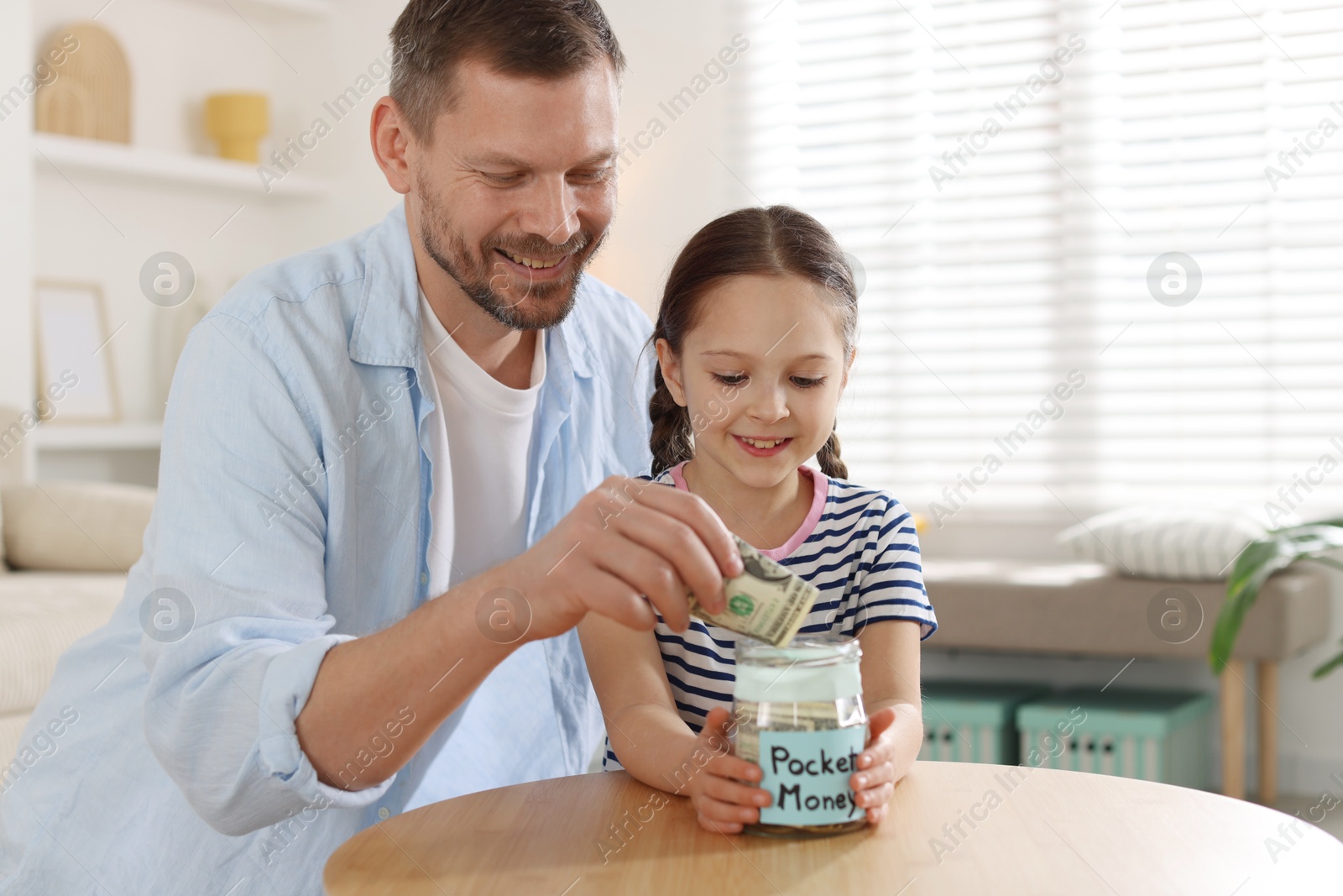Father and daughter putting pocket money into jar at table indoors Photo of Father and daughter putting pocket money into jar at table indoors